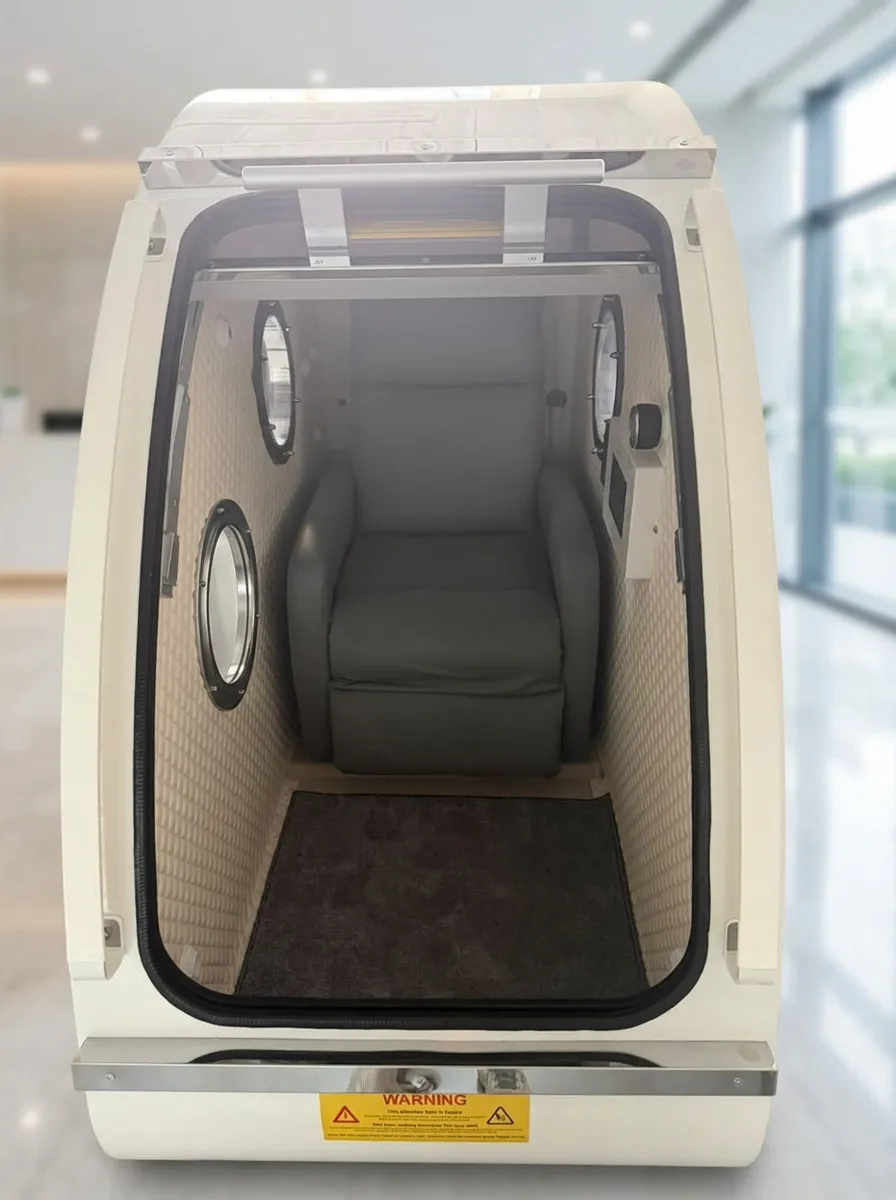 Front view of the open white hard-shell chamber with a grey leather recliner seat and extended footrest inside, quilted cream padded walls, three round viewing windows, and a wall-mounted touchscreen control panel